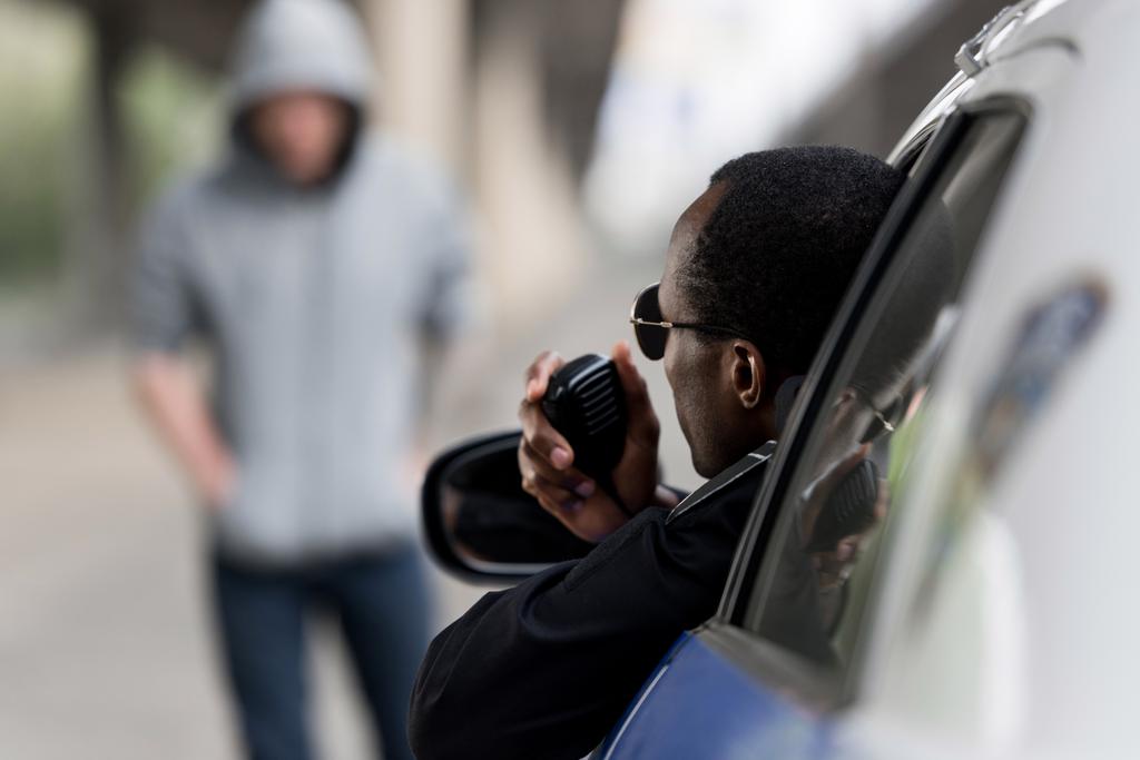 Полицейский смотрит на молодого человека в капюшоне на улице из окна и разговаривает по радио
 - Фото, изображение