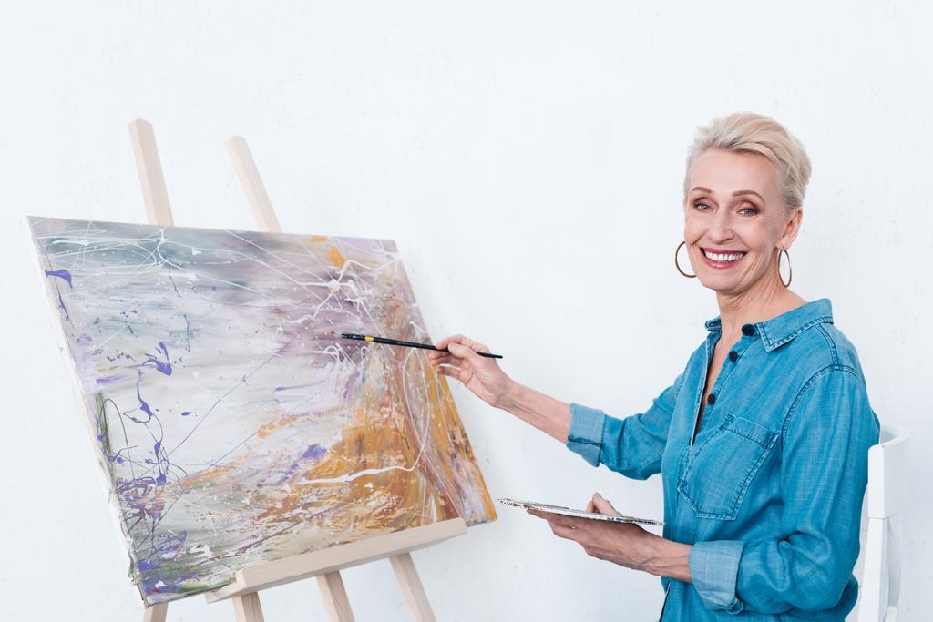 senior cheerful woman painting with palette on easel in workshop - Photo, Image