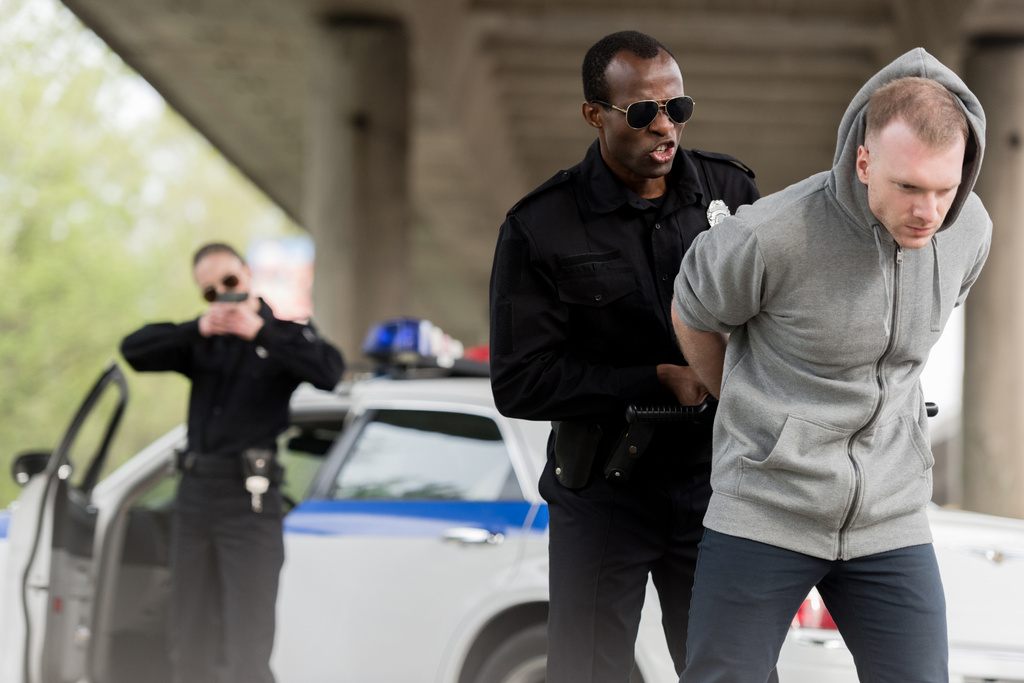 сердитий афроамериканський поліцейський заарештовує молодого чоловіка, а жінка-партнер прикриває його пістолетом
  - Фото, зображення