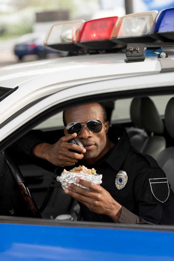 policier afro-américain tenant un hamburger et parlant sur la radio portable dans la voiture
  - Photo, image