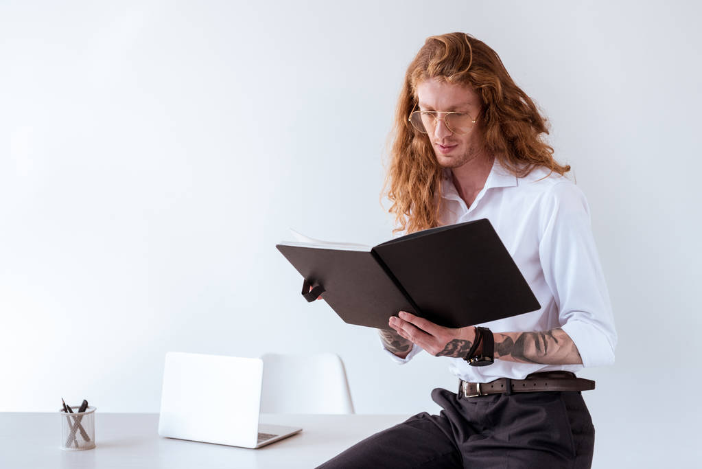 homem de negócios tatuado elegante com cabelo encaracolado sentado na mesa e ler documentos no local de trabalho
 - Foto, Imagem