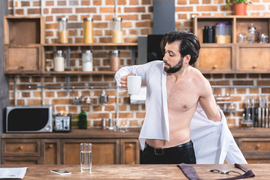 handsome loner businessman wearing shirt and holding cup of coffee at kitchen - Photo, Image