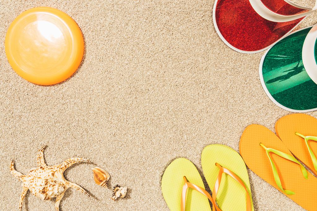 flat lay with flying disk, colorful caps, flip flops and seashells on sand - Photo, Image