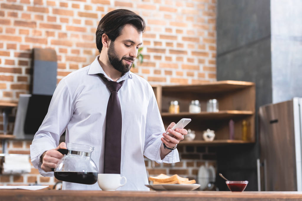 guapo hombre de negocios solitario sosteniendo la olla con café y teléfono inteligente en la cocina
 - Foto, imagen