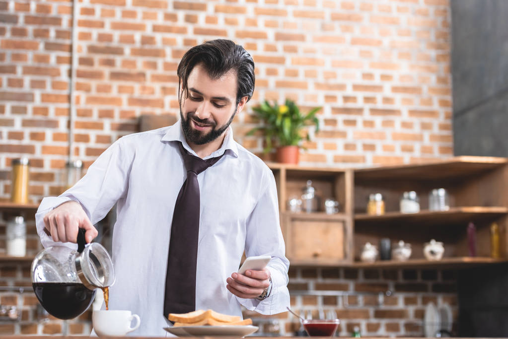 komea yksinäinen liikemies kaatamalla kahvia kuppiin ja pitämällä älypuhelin keittiössä
 - Valokuva, kuva
