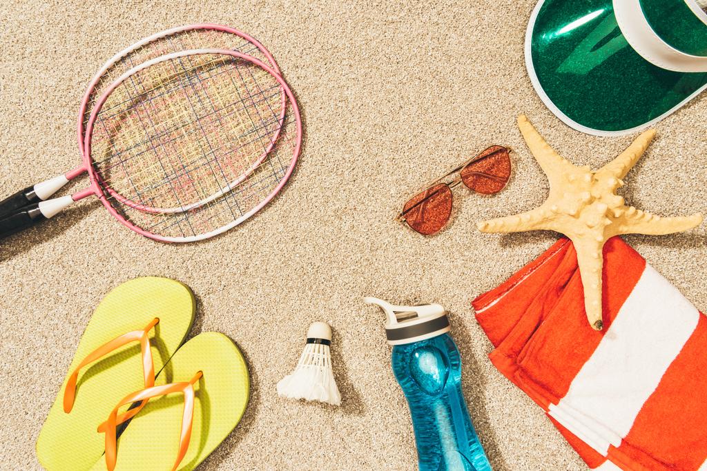 Blick von oben auf Badminton-Ausrüstung, Sonnenbrille, Flip Flops, Mütze und Handtuch auf Sand - Foto, Bild