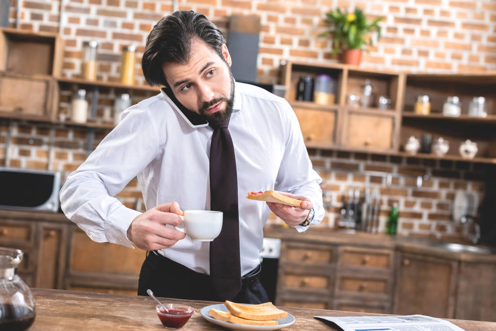 loner businessman talking by smartphone while having breakfast at kitchen - Photo, Image