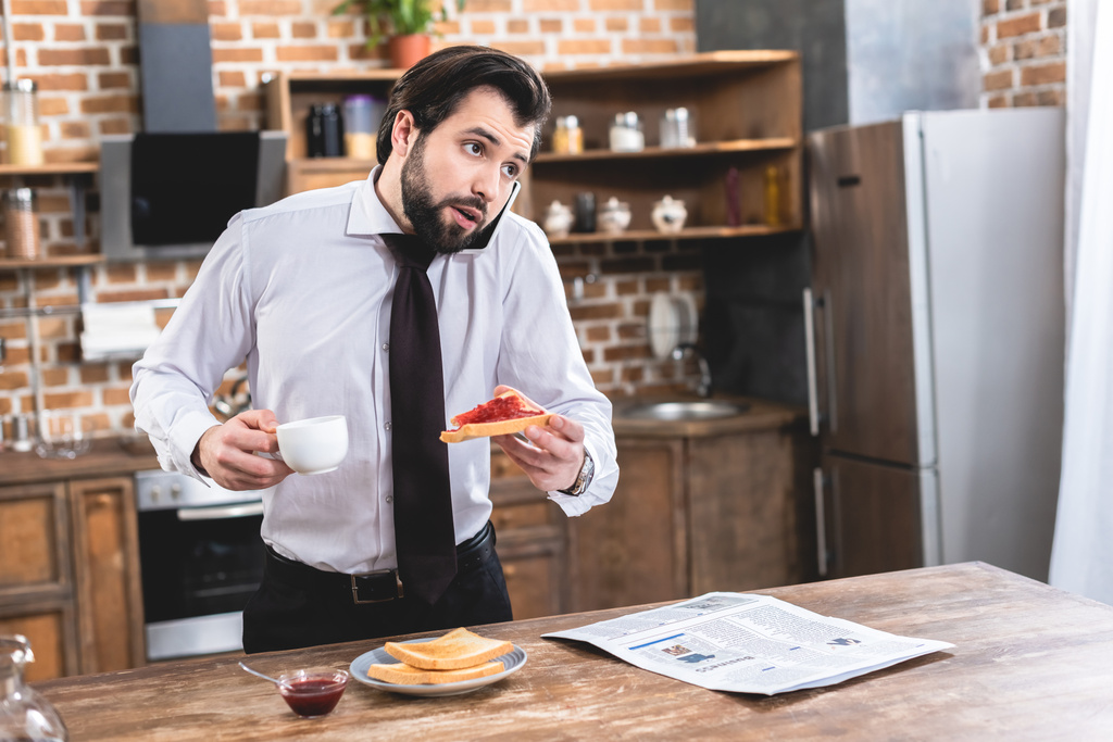 samotnik przystojny biznesmen rozmawia przez smartfona, jedząc śniadanie w kuchni - Zdjęcie, obraz
