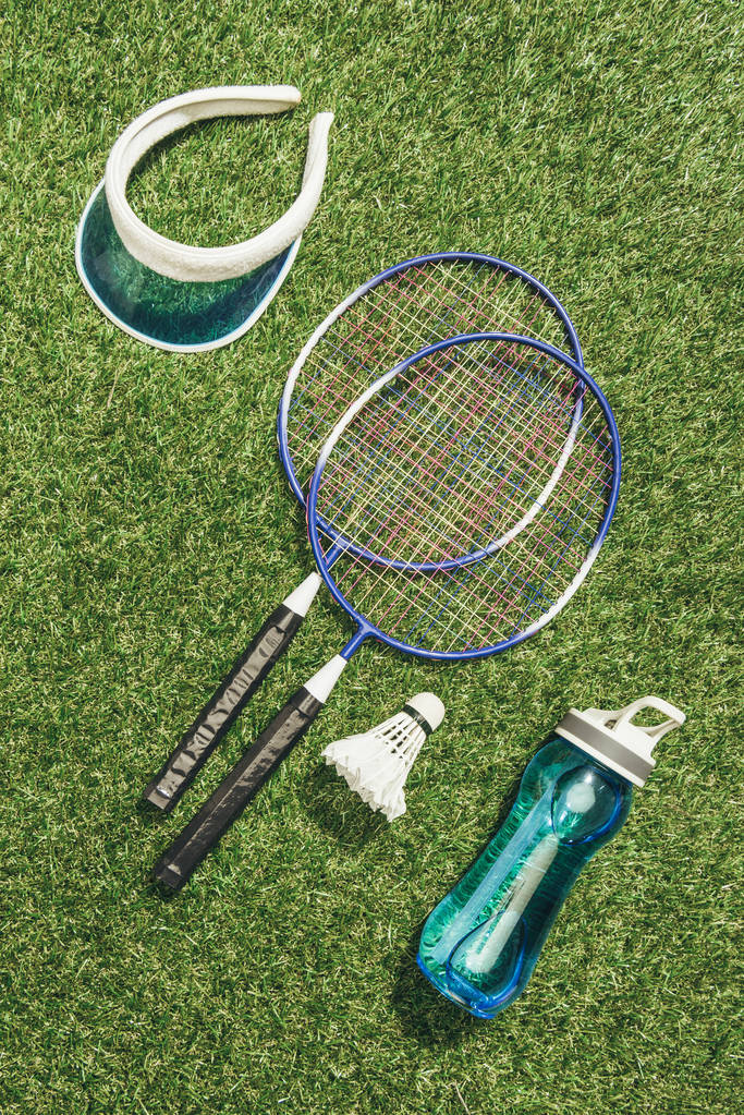 Blick von oben auf Badminton-Ausrüstung, Wasserflasche und Kappe auf grünem Gras - Foto, Bild
