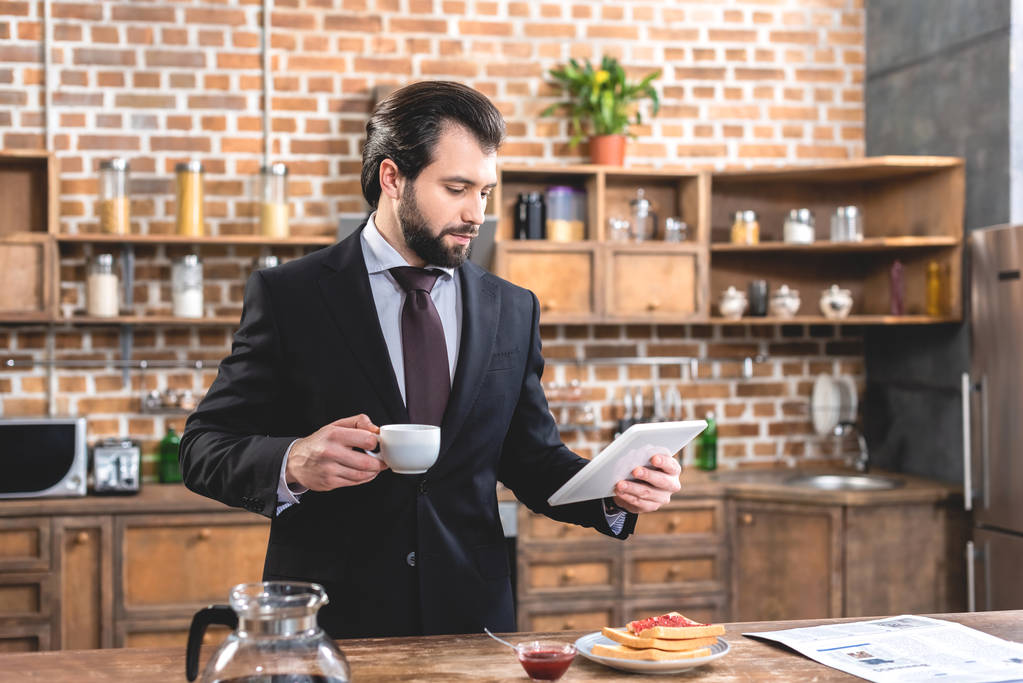 komea yksinäinen liikemies tilalla tabletti ja kuppi kahvia keittiössä
 - Valokuva, kuva