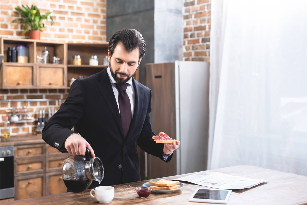 όμορφος μοναχικός επιχειρηματίας έκχυση καφέ και κρατώντας τοστ στην κουζίνα - Φωτογραφία, εικόνα