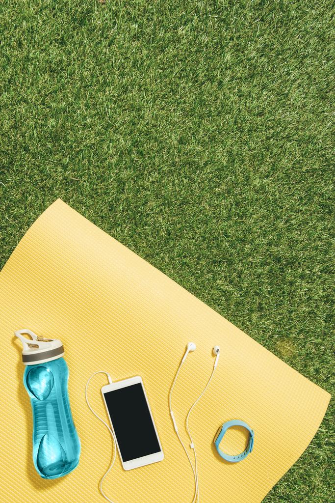 flache Liege mit Wasserflasche, Uhr, Smartphone und Kopfhörer auf gelber Matte auf grünem Rasen - Foto, Bild