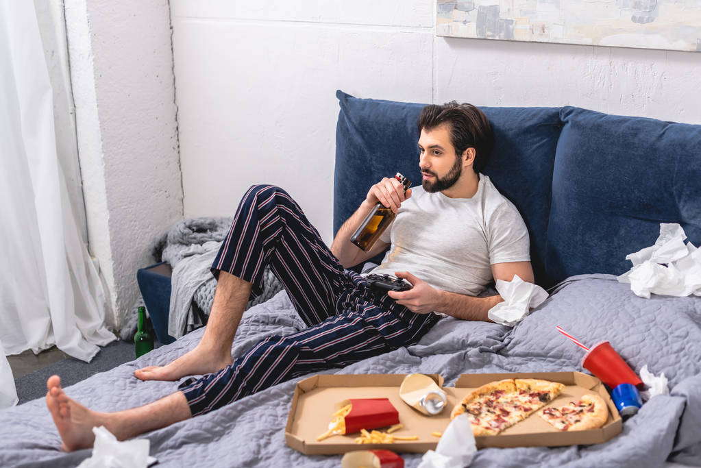 masculino solitário jogar vídeo game e beber cerveja na cama em casa
 - Foto, Imagem