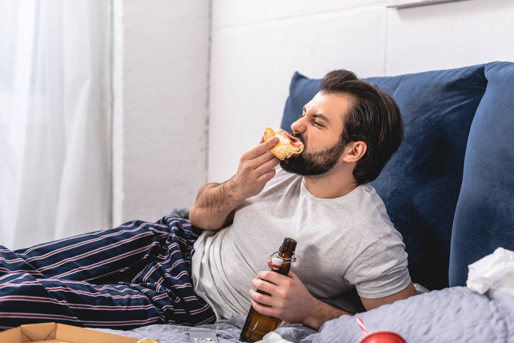 knappe eenling eten van pizza en flesje bier houden in slaapkamer - Foto, afbeelding
