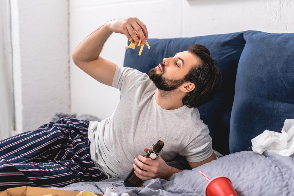 guapo solitario comiendo papas fritas y sosteniendo botella de cerveza en el dormitorio
 - Foto, Imagen
