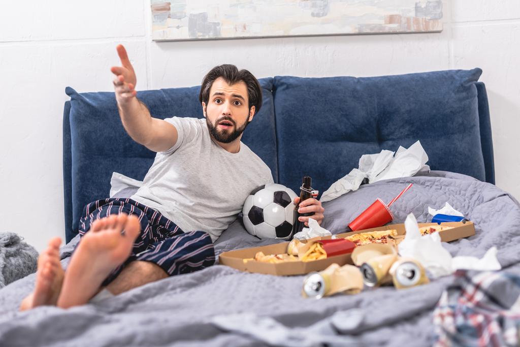 guapo solitario gesto mientras mira partido de fútbol con cerveza en el dormitorio
 - Foto, imagen