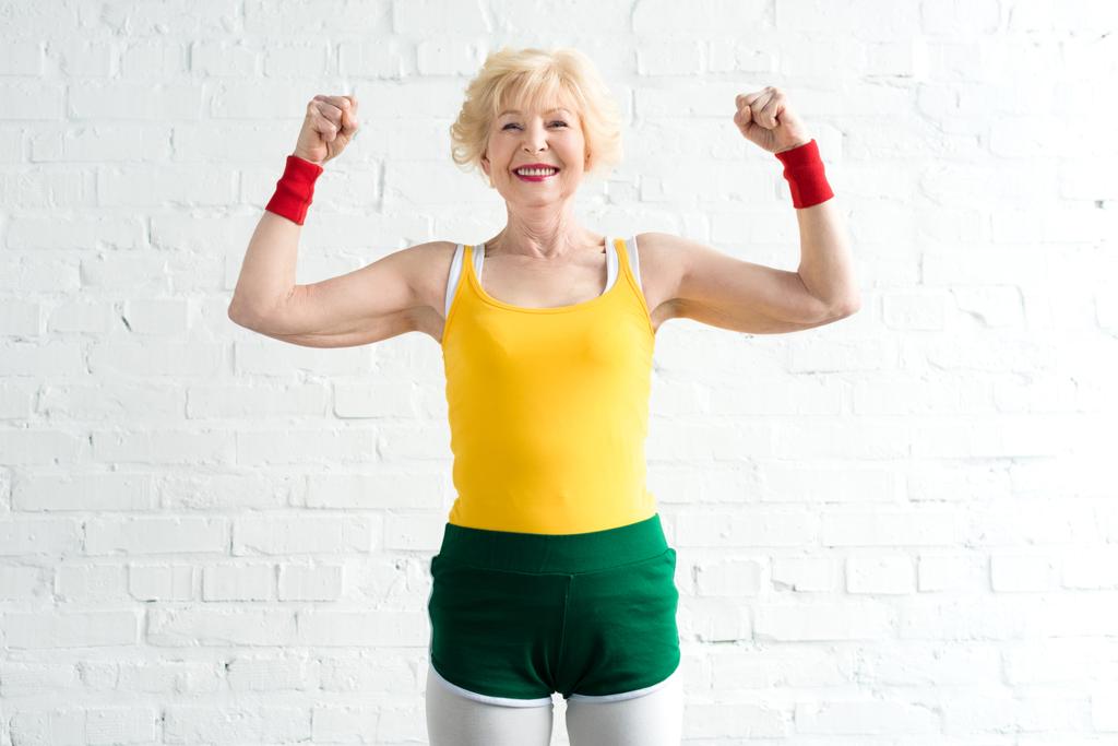 Gelukkig senior vrouw in sportkleding tonen van spieren en glimlachend op camera  - Foto, afbeelding