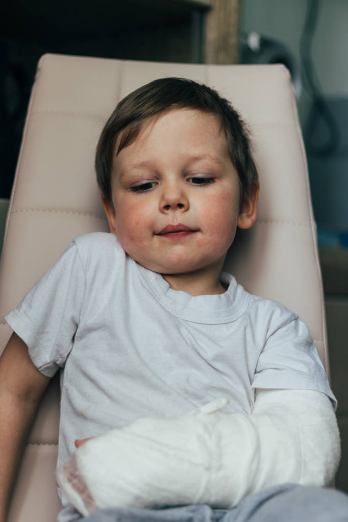 un niño pequeño está sentado en una silla con un brazo roto, en un yeso. hospital. medicina. peligro. dolor. para romper extremidades. violencia
. - Foto, Imagen