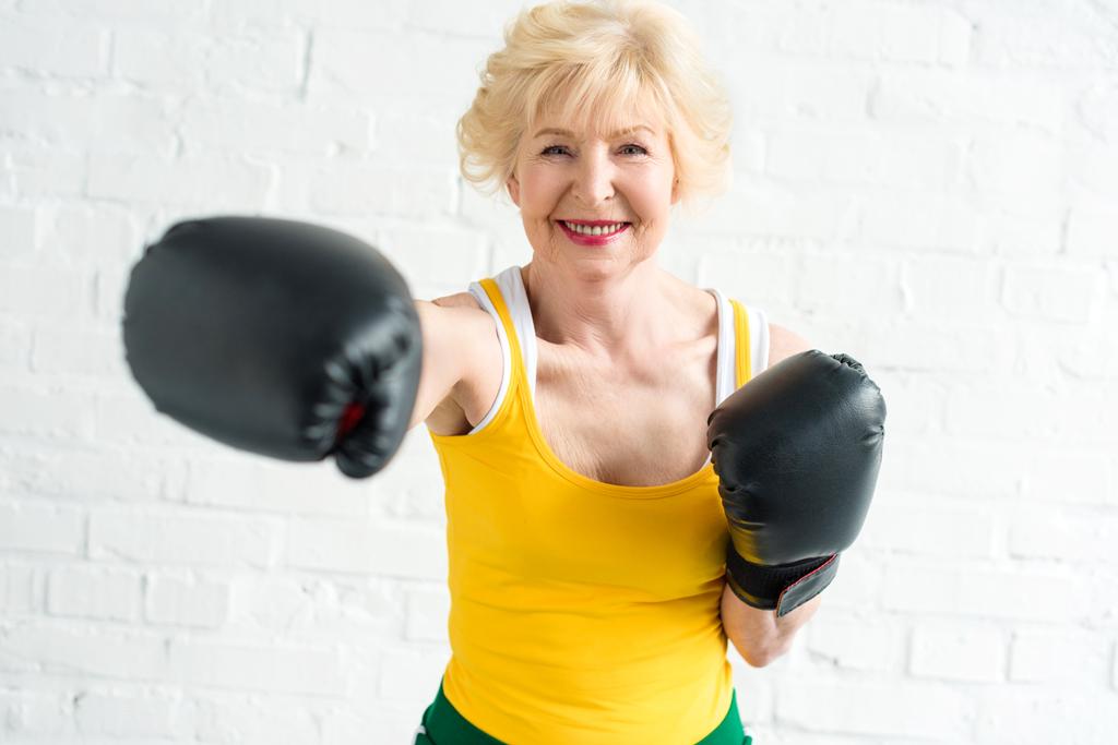 feliz mujer mayor en el entrenamiento de guantes de boxeo y sonriendo a la cámara
 - Foto, Imagen