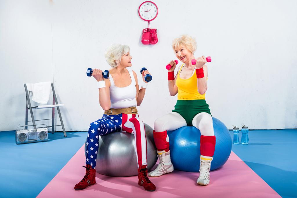 mulheres idosas em treinamento sportswear com halteres enquanto sentado em bolas de fitness e sorrindo uns aos outros
 - Foto, Imagem