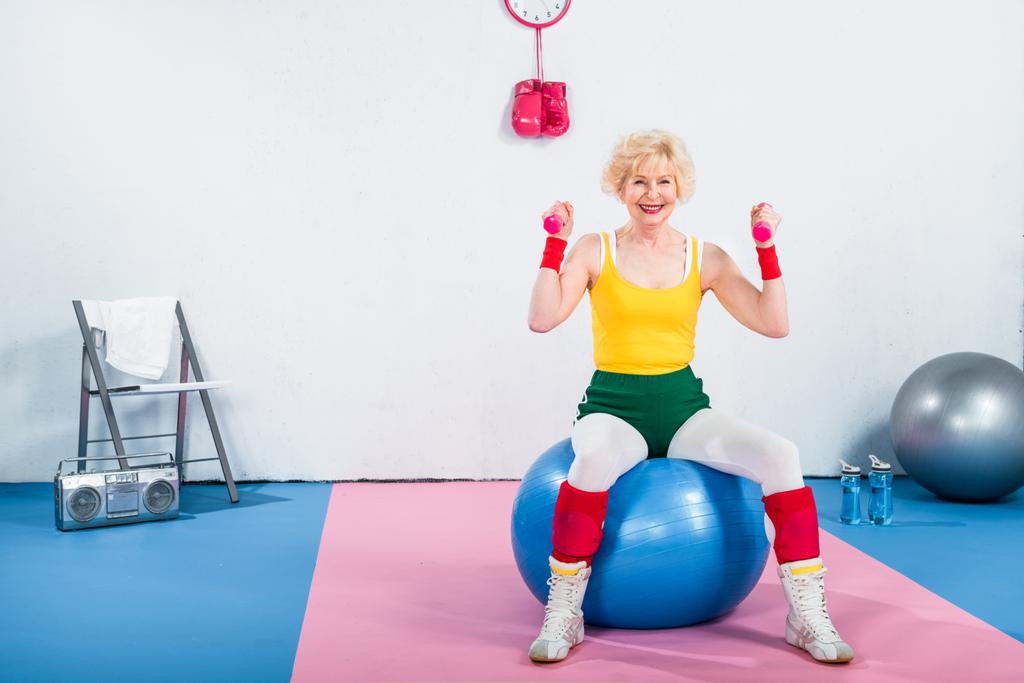 femme âgée sportive souriante assise sur un ballon de fitness et faisant de l'exercice avec des haltères
  - Photo, image