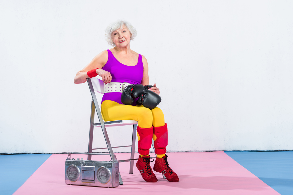 heureuse femme âgée sportive avec des gants de boxe assis et souriant à la caméra
  - Photo, image