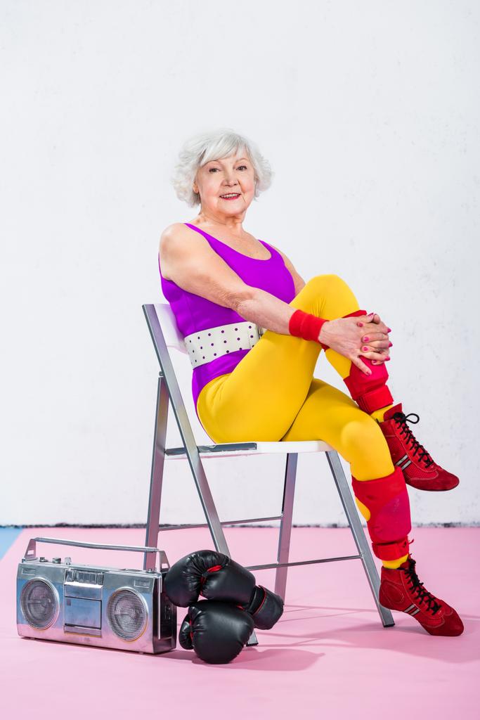 Glückliche Seniorin sitzt neben Tonbandgerät mit Boxhandschuhen und lächelt in die Kamera - Foto, Bild