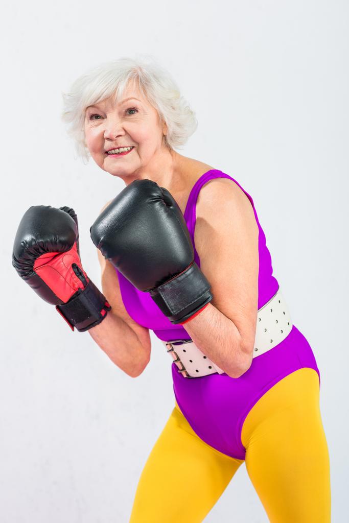 Портрет красивой спортивной пожилой женщины в боксёрских перчатках, улыбающейся в камеру, изолированную на сером
 - Фото, изображение