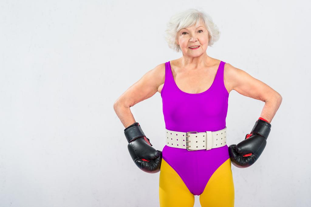 счастливая старшая спортсменка в боксёрских перчатках, стоящая с руками на талии и улыбающаяся в камеру, изолированную на сером
 - Фото, изображение