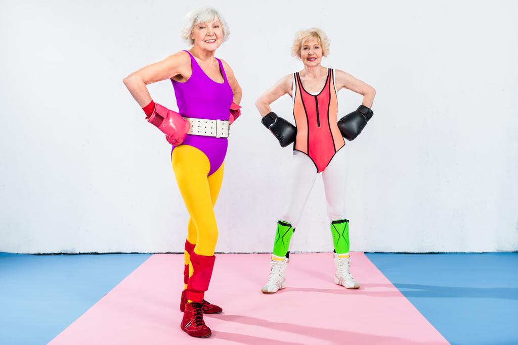 volle lengte weergave van sportieve senior dames in bokshandschoenen permanent met handen op taille en lachend op camera - Foto, afbeelding