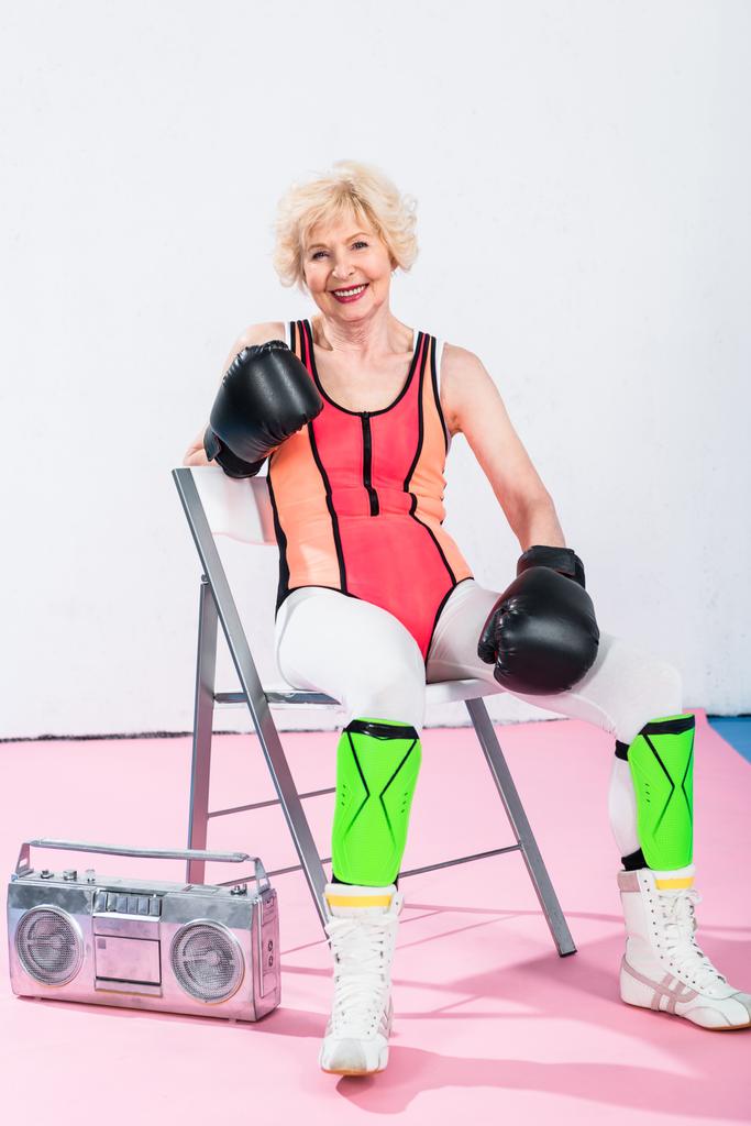 sporty senior woman in boxing gloves sitting with tape recorder and smiling at camera - Photo, Image