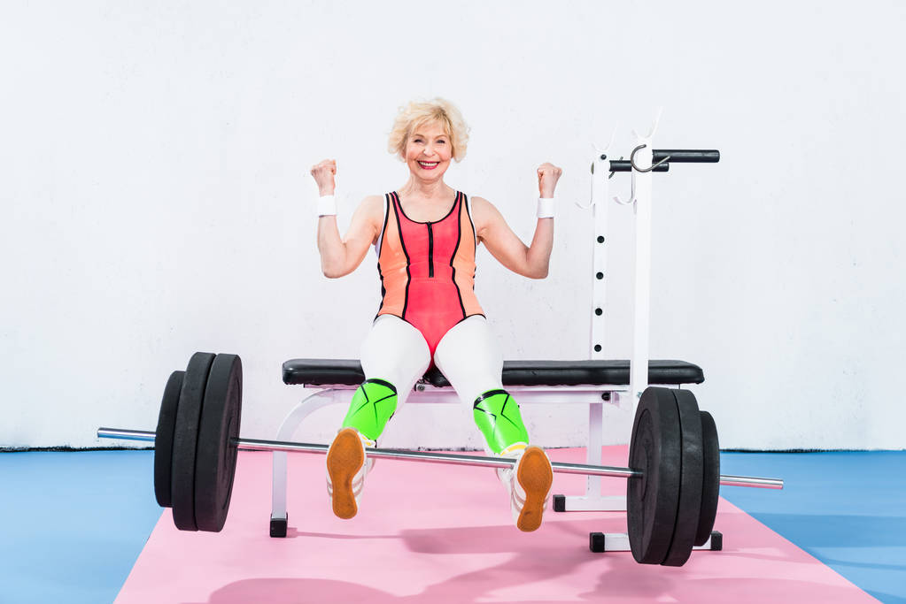 ευτυχής ανώτερος αθλήτρια άρση της barbell με τα πόδια και να χαμογελά στη φωτογραφική μηχανή - Φωτογραφία, εικόνα