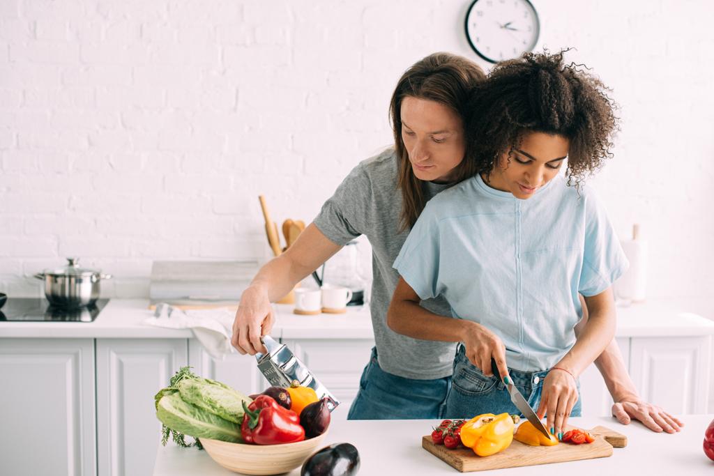 jovem mulher cortando pimentão e namorado olhando ralador na cozinha
  - Foto, Imagem