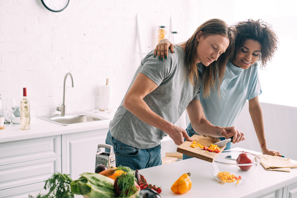 smiling african american woman embracing boyfriend while he cooking salad at kitchen - Photo, Image