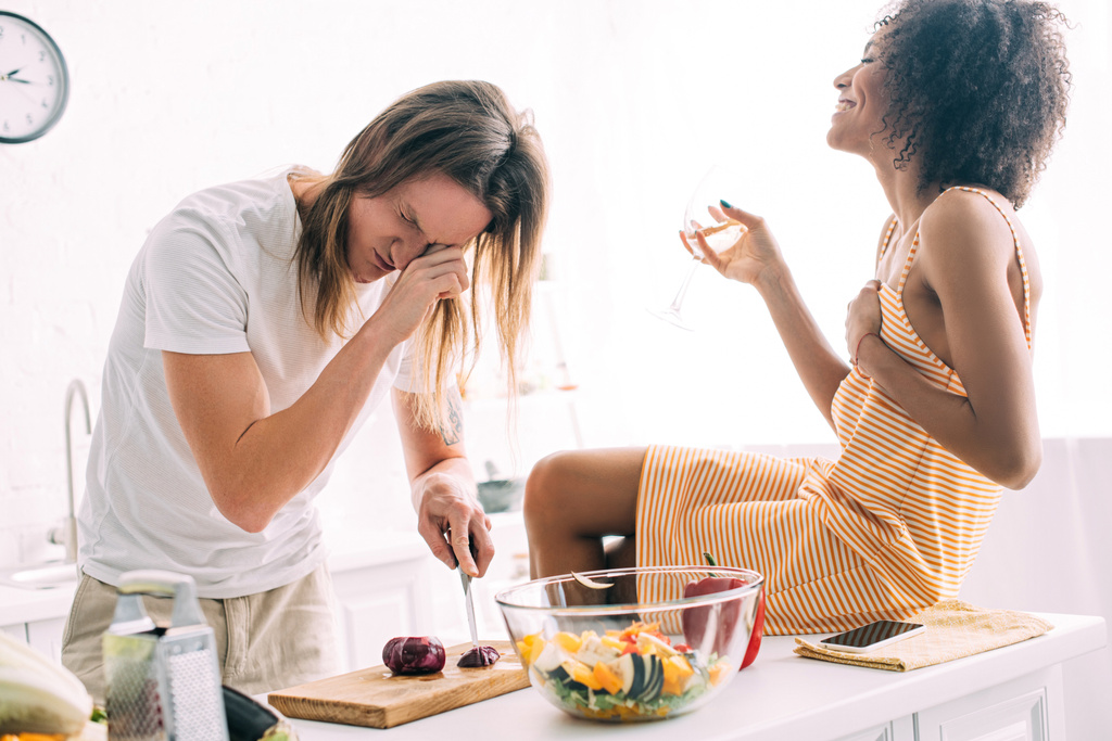 s úsměvem afroamerické ženy se víno sedí na kuchyň, zatímco její přítel řezání cibule a pláč v kuchyni - Fotografie, Obrázek