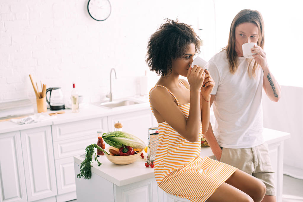 Sex tussen verschillendre rassen jong koppel koffie drinken uit witte bekers op keuken  - Foto, afbeelding