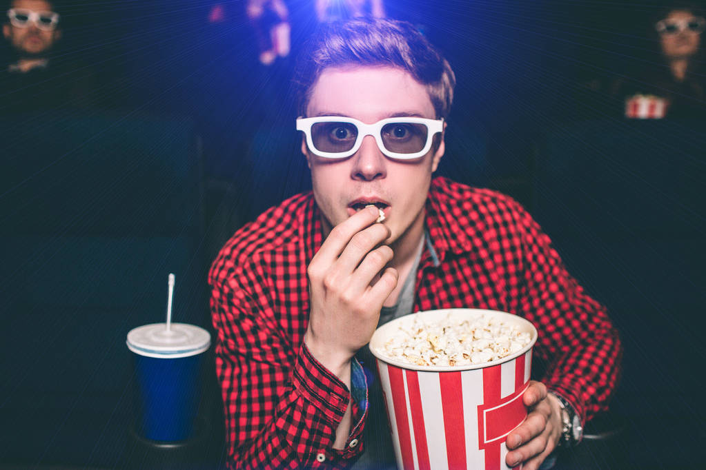 Портрет парня, сидящего в кресле в кинозале и поедающего попкорн. Он выглядит очень напряженным. Парень носит специальные очки для просмотра 3D фильмов. Также он ест попкорн из корзины
. - Фото, изображение