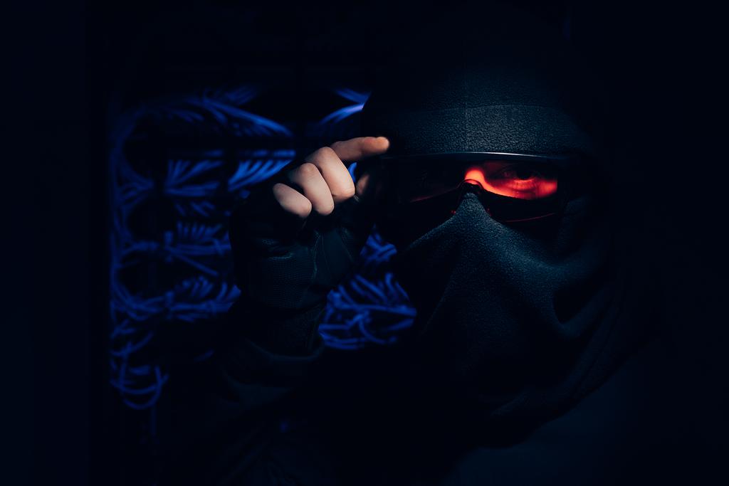 obscured view of hacker in eyeglasses and mask looking at camera with cables on background - Photo, Image