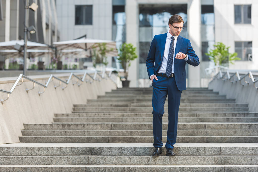 csinos fiatal üzletember állva lépcsők közelében üzleti épület és látszó-on néz elegáns öltöny - Fotó, kép