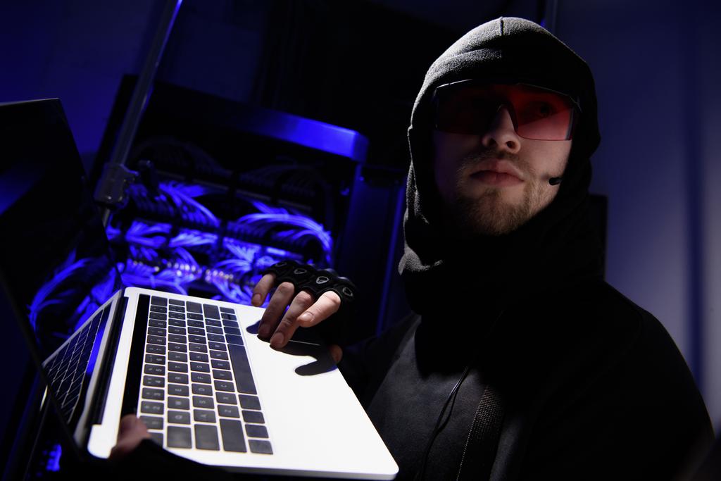 Seitenansicht des Hackers in Brille mit Laptop, der mit Kabeln im Hintergrund wegschaut - Foto, Bild