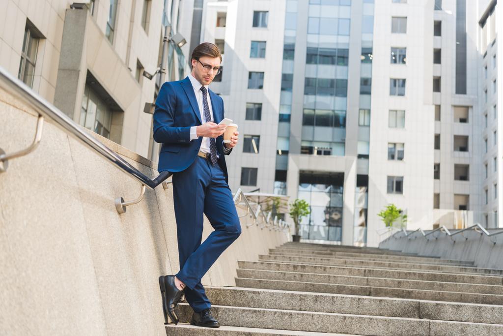 attractive young businessman in stylish suit with paper cup of coffee using smartphone on stairs near business building - Photo, Image