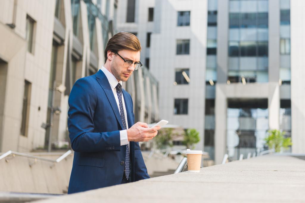 vonzó fiatal üzletember, elegáns színű papír csésze kávé használ smartphone üzleti épület közelében - Fotó, kép