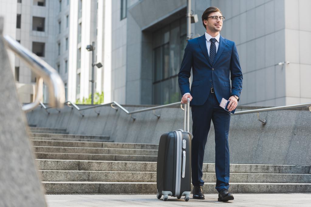 successful businessman in stylish suit with luggage and flight tickets going down stairs in business district - Photo, Image