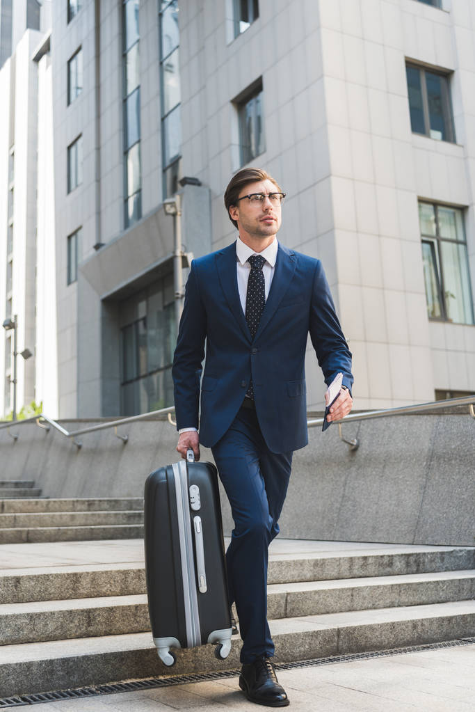 bonito jovem empresário em terno elegante com bagagem e bilhetes de avião descendo escadas no distrito de negócios
 - Foto, Imagem