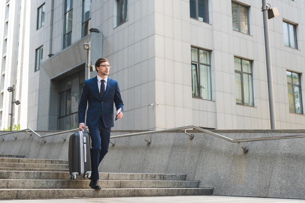 бизнесмен в стильном костюме с багажом и авиабилетами спускается по лестнице в деловой район
 - Фото, изображение