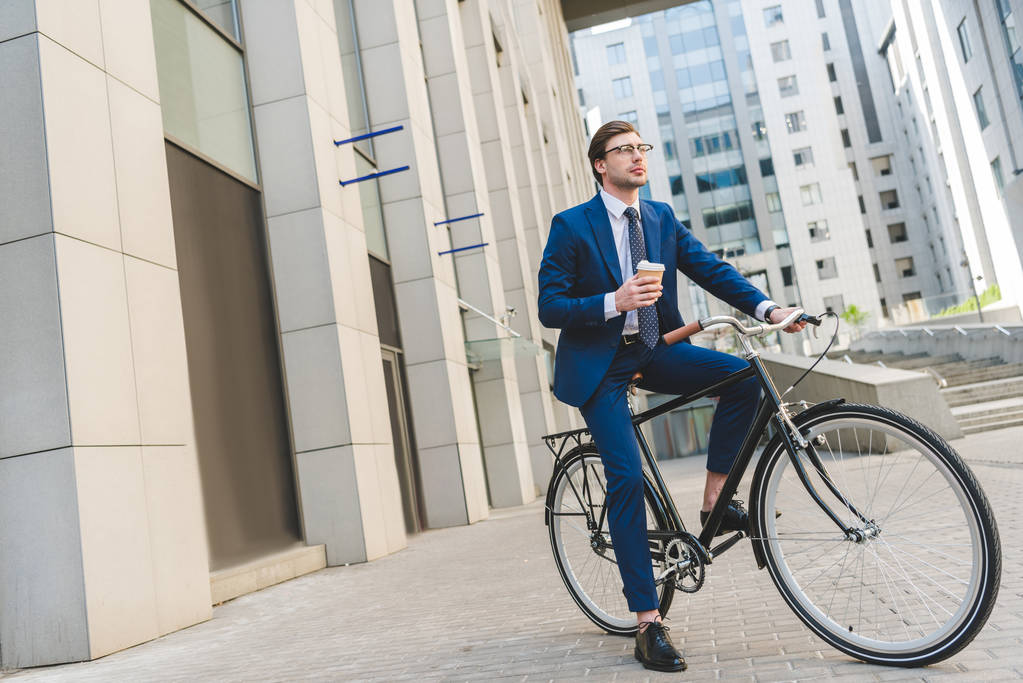 schöner junger Geschäftsmann im schicken Anzug mit Kaffee zum Sitzen auf einem Oldtimer-Fahrrad - Foto, Bild
