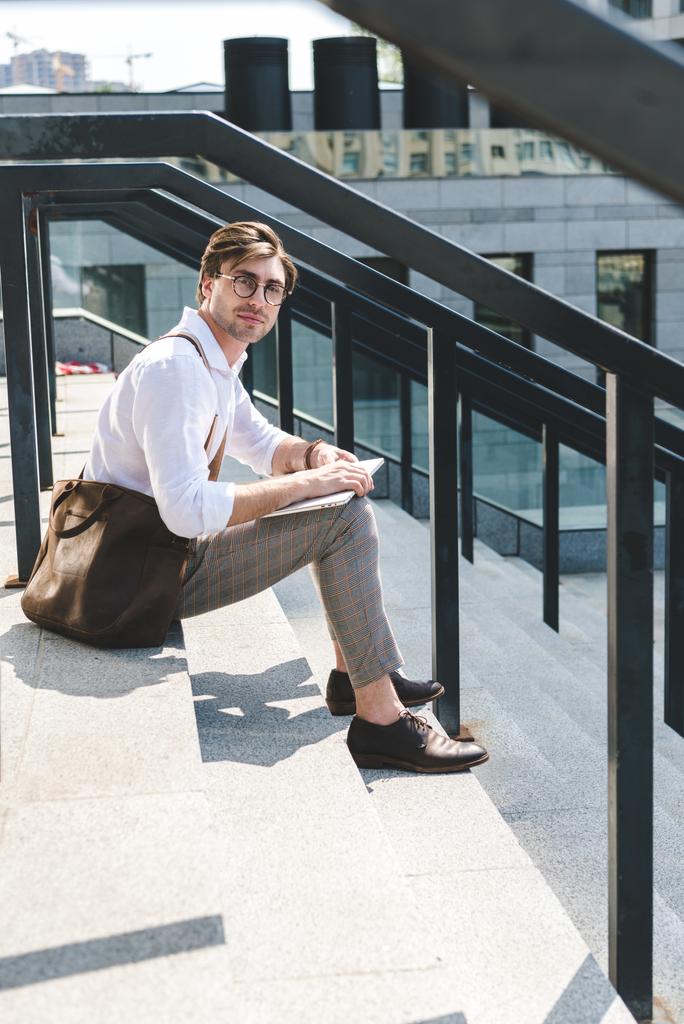 красивый молодой человек с ноутбуком смотрит в камеру на лестнице на городской улице
 - Фото, изображение
