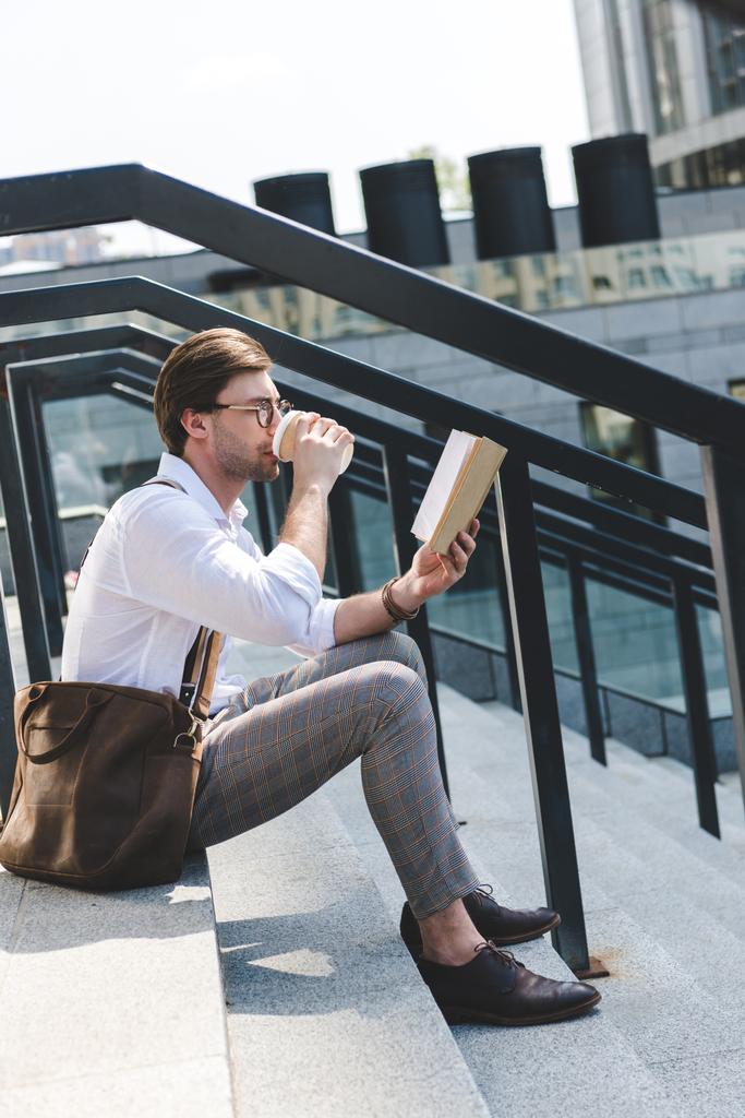 pohledný mladý muž popíjel kávu z papíru pohár a čtení knihy na schodech na ulici - Fotografie, Obrázek