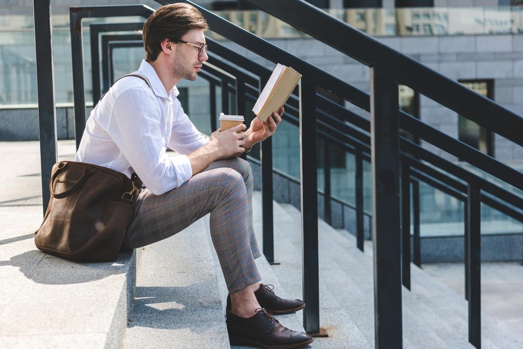 vista laterale di bel giovane uomo con carta tazza di caffè lettura libro sulle scale sulla strada della città
 - Foto, immagini