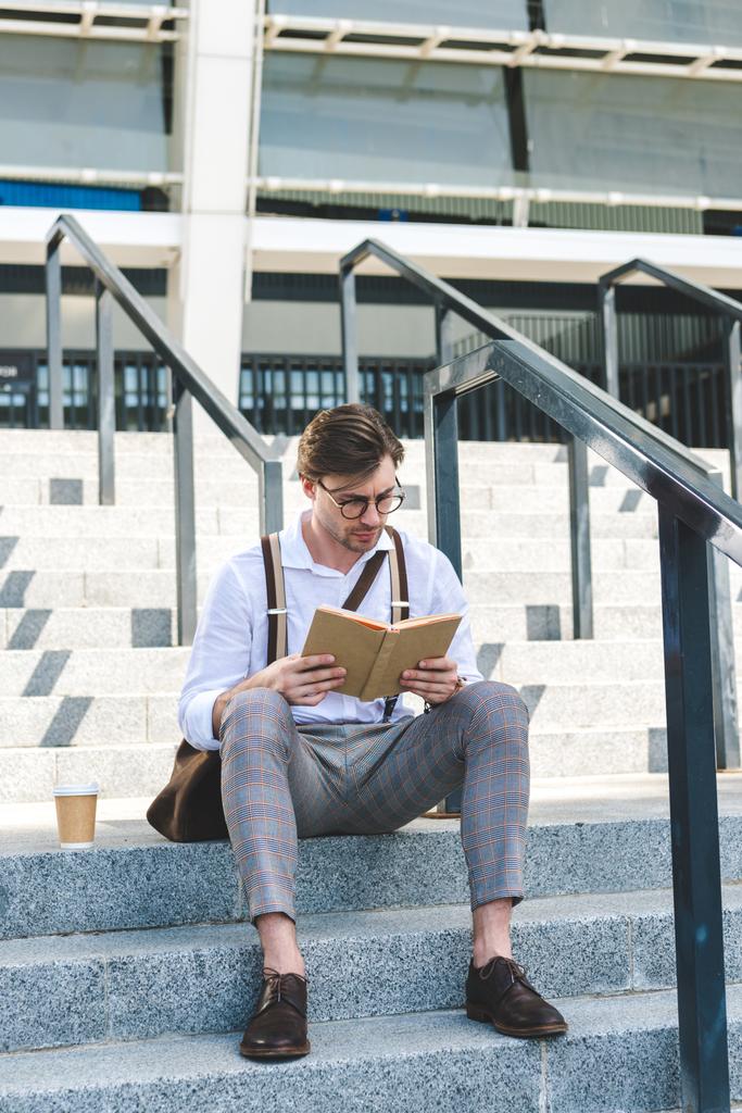 ориентированный молодой человек с бумажной чашкой кофе чтение книги на лестнице на городской улице
 - Фото, изображение